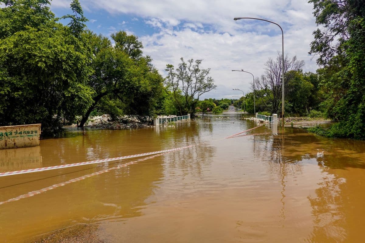 A flooded road with a submerged bridge, surrounded by trees, under a cloudy sky.